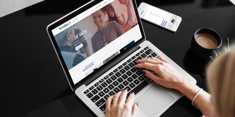 Mulher olha para tela do notebook que contém a página inicial do site da Fundepar, sob a mesa está um celular que também mostra o site na versão mobile